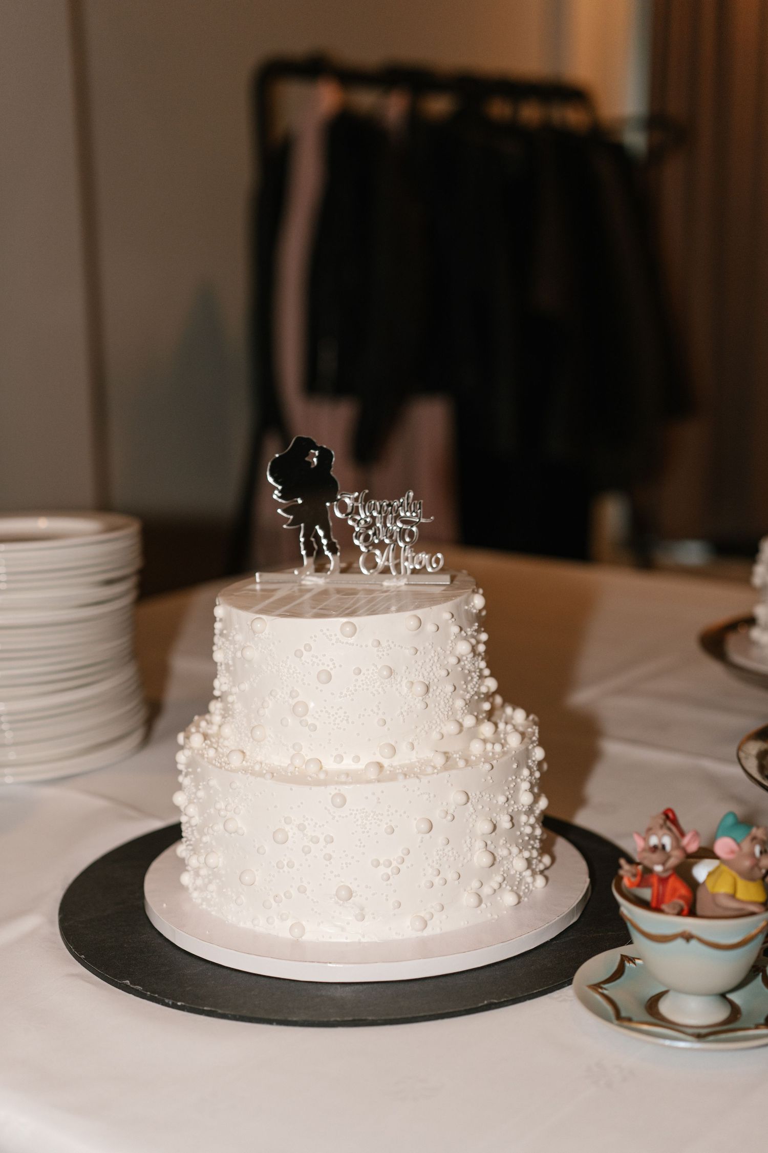 White wedding cake displayed on black plate with decorative tea set nearby.