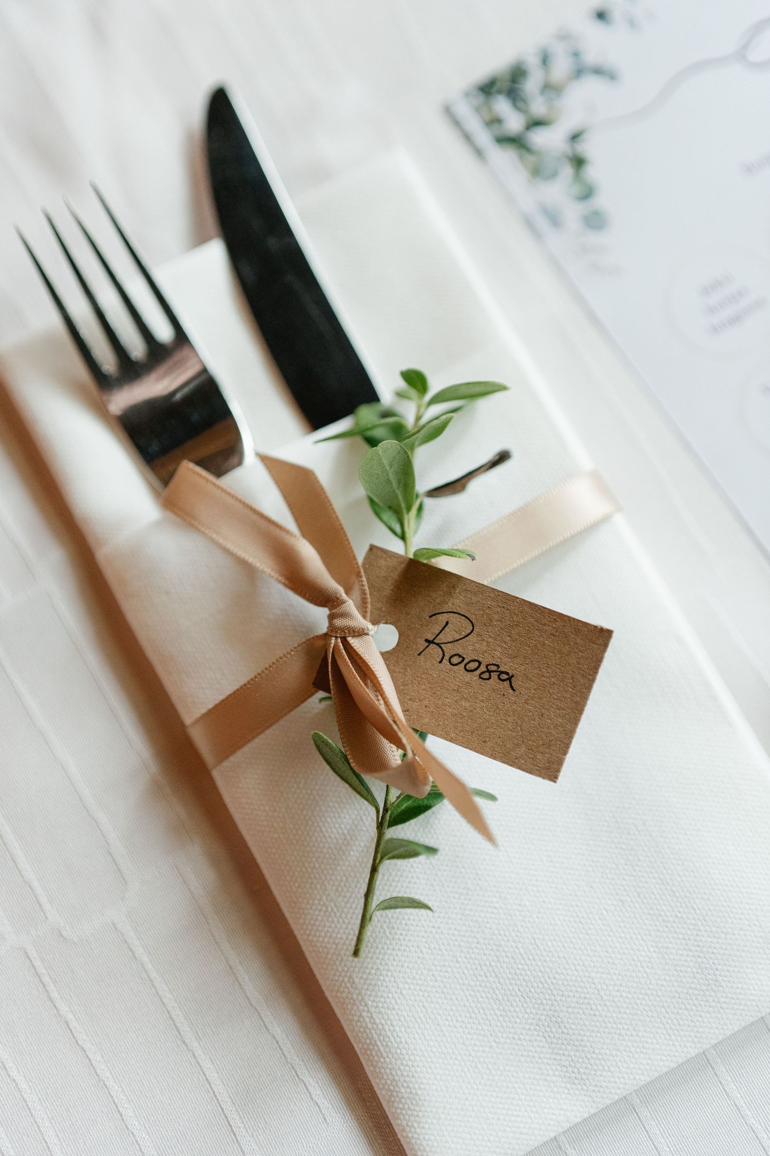 Elegant place setting with white napkin, silverware, and fresh greenery tied with kraft paper name tag.