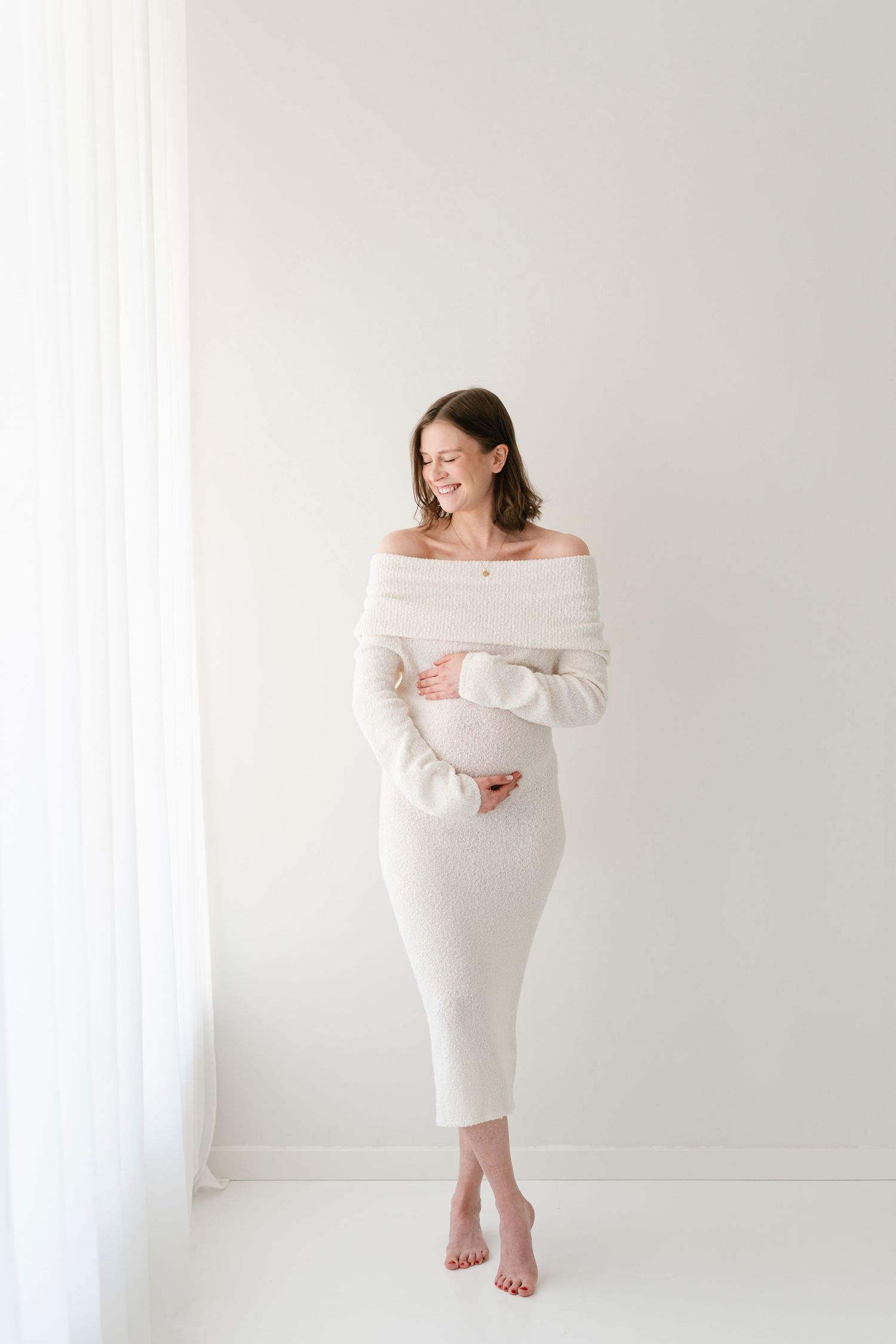 Expectant mother in flowing white dress poses gracefully in bright minimalist studio setting.