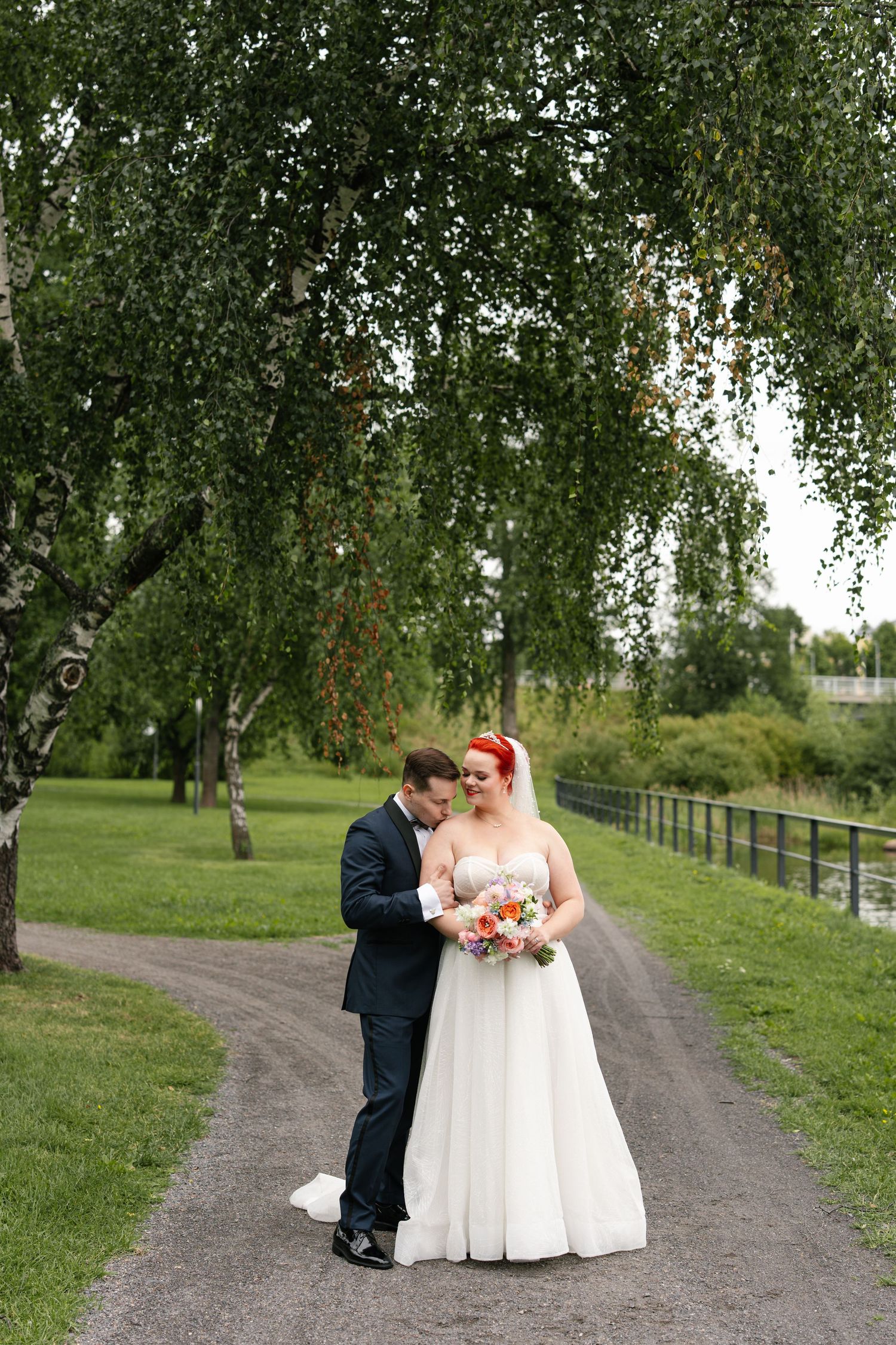 Wedding couple poses on tree-lined path with natural outdoor backdrop.