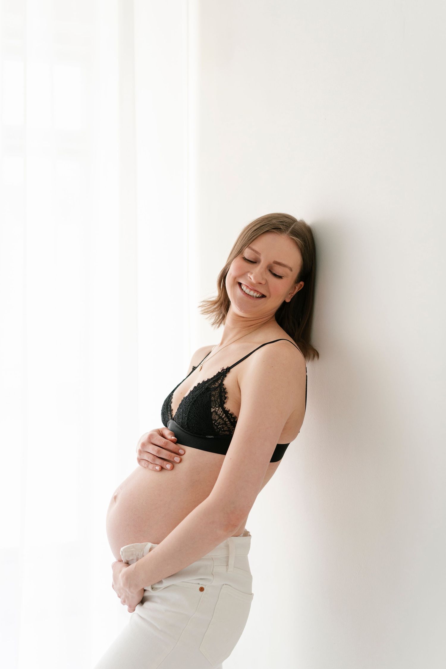 Joyful maternity portraits showing woman in black bra and white pants against white wall.