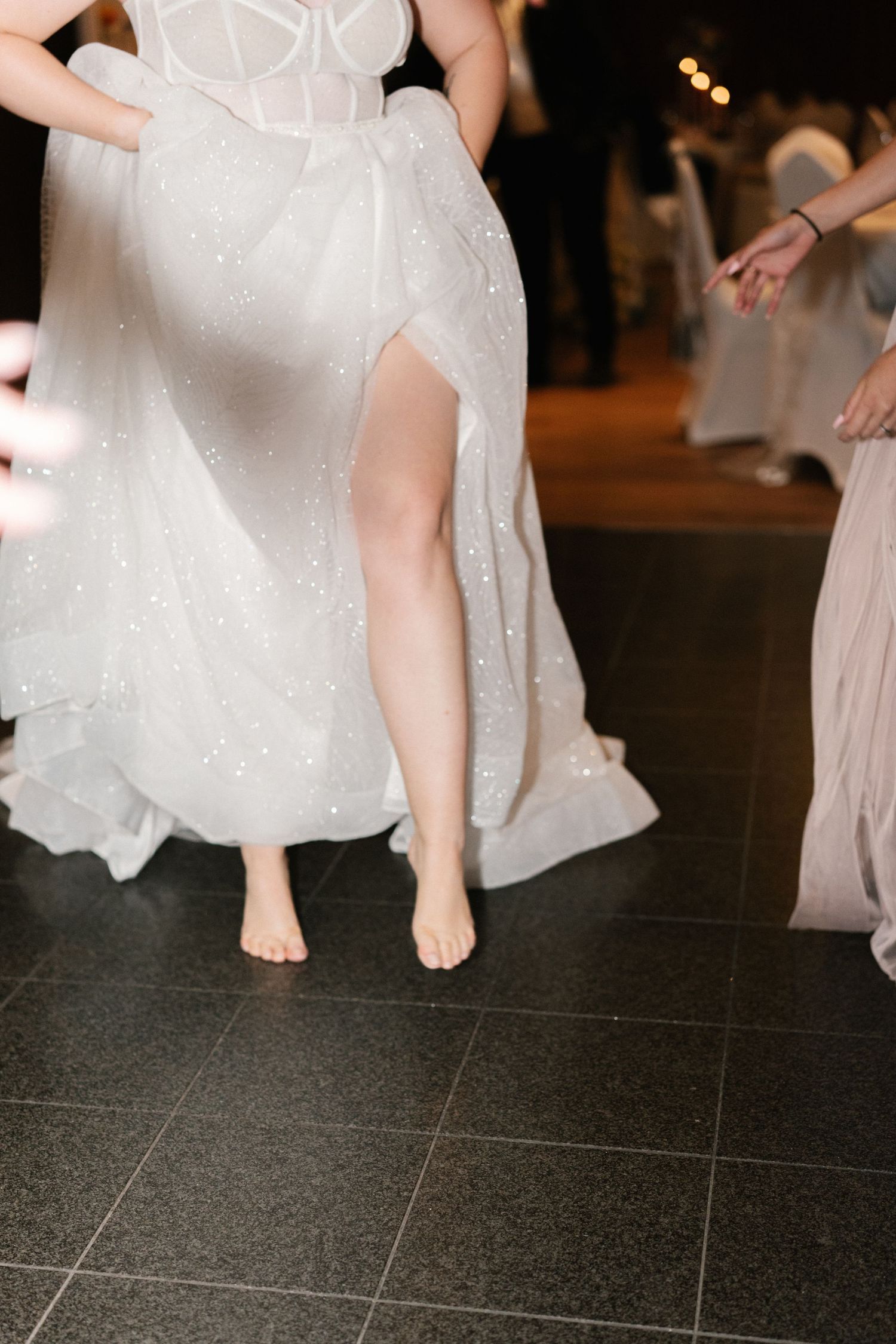 Close-up of a bride's bare feet under her white wedding dress on a dark dance floor.