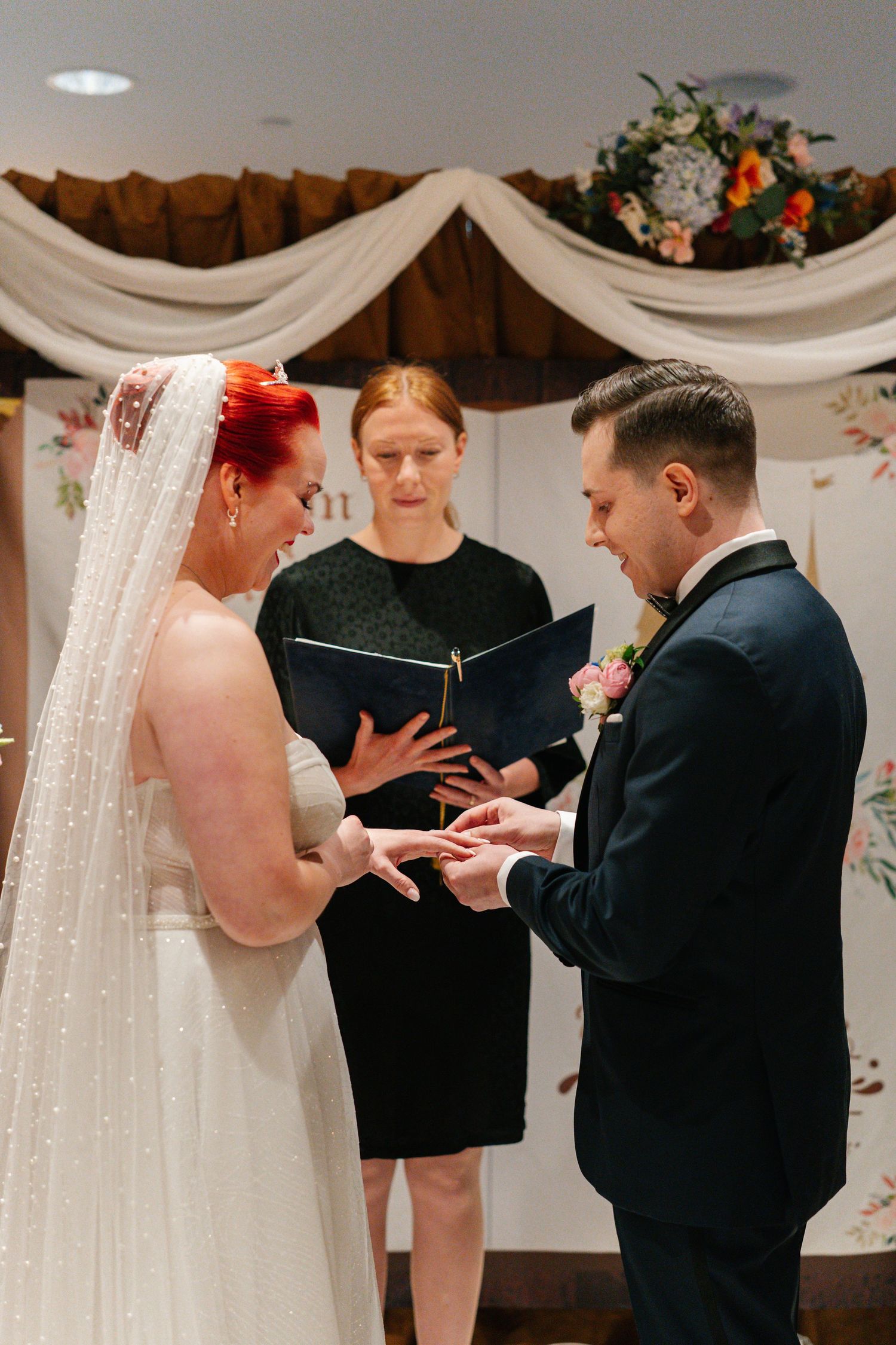 A couple exchanges rings during an intimate wedding ceremony under white draped fabric and floral decorations.