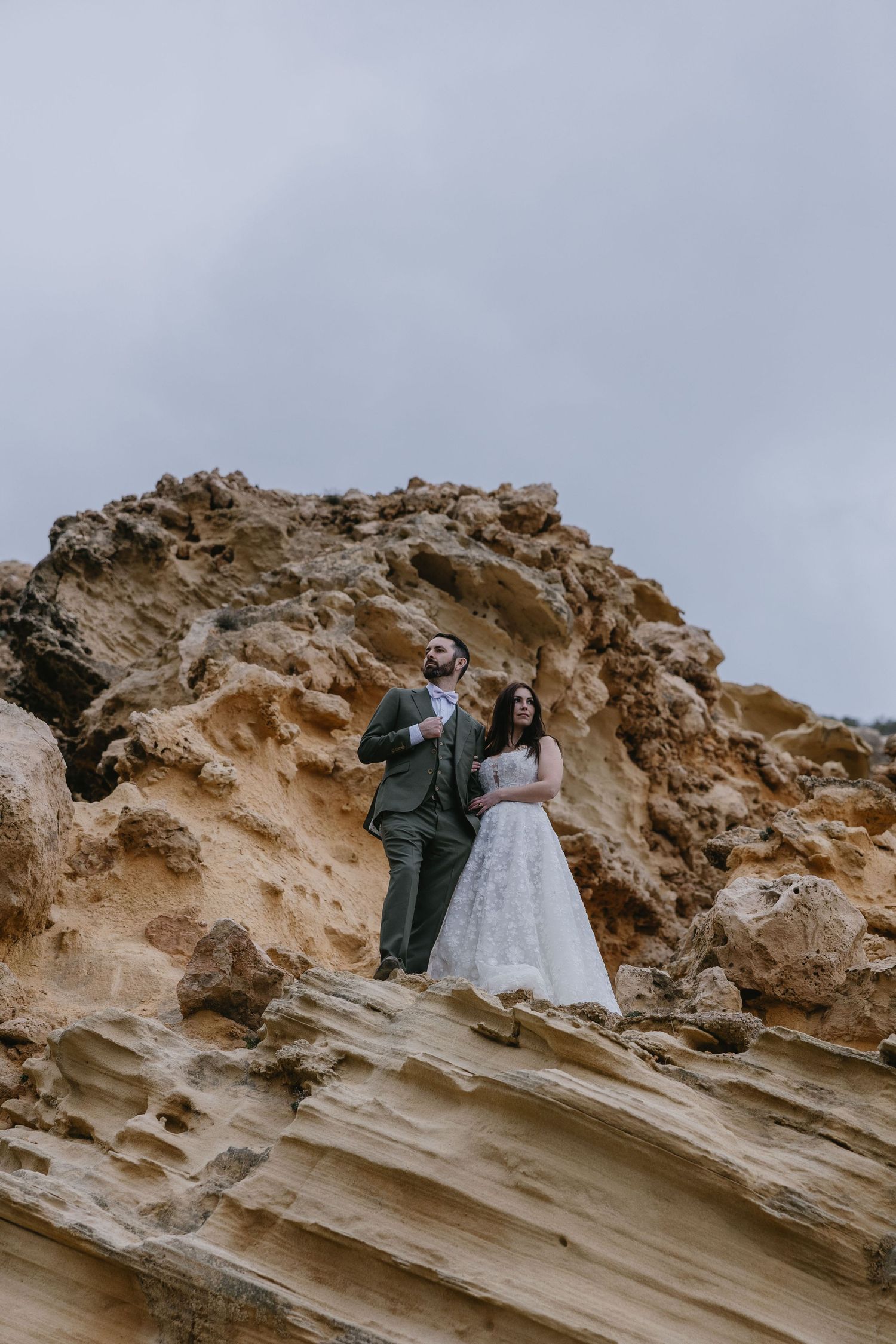Elopement couple standing atop a dramatic rocky cliff, embracing against an overcast sky for an adventurous and cinematic wedding portrait.