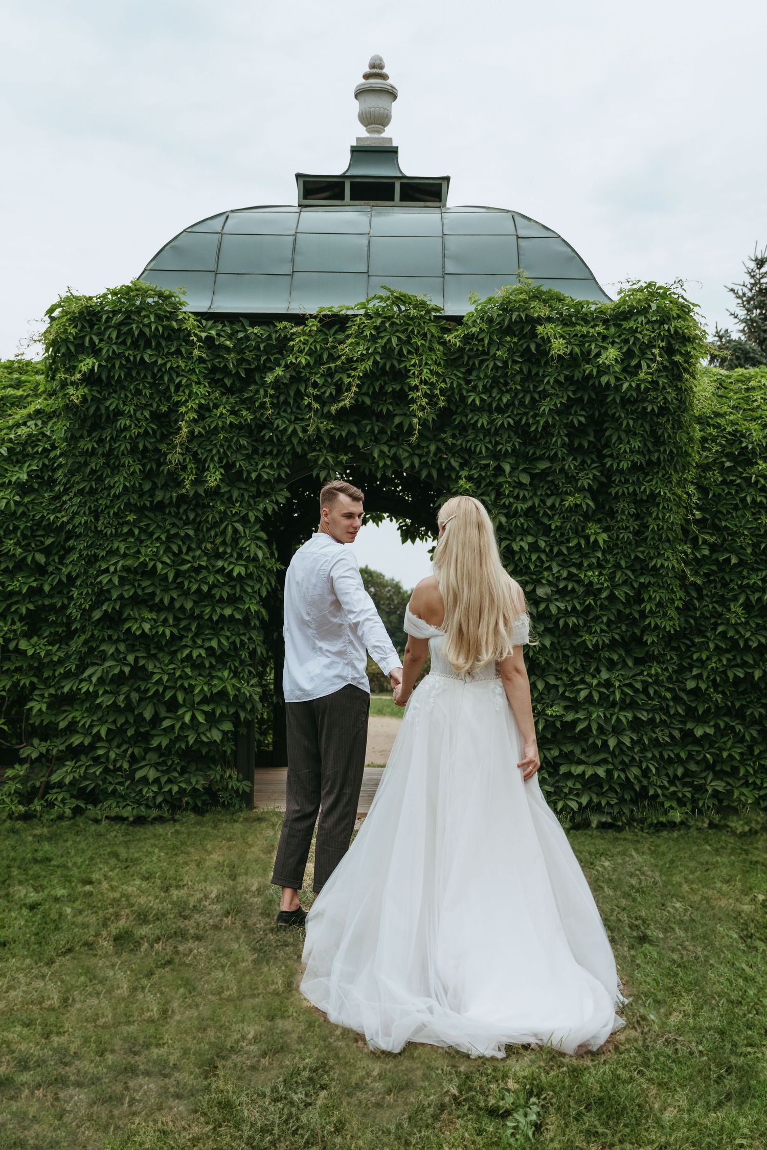 A bride and groom share a quiet moment in a lush villa garden, framed by an ivy-covered gazebo with a domed roof - elegant European wedding.