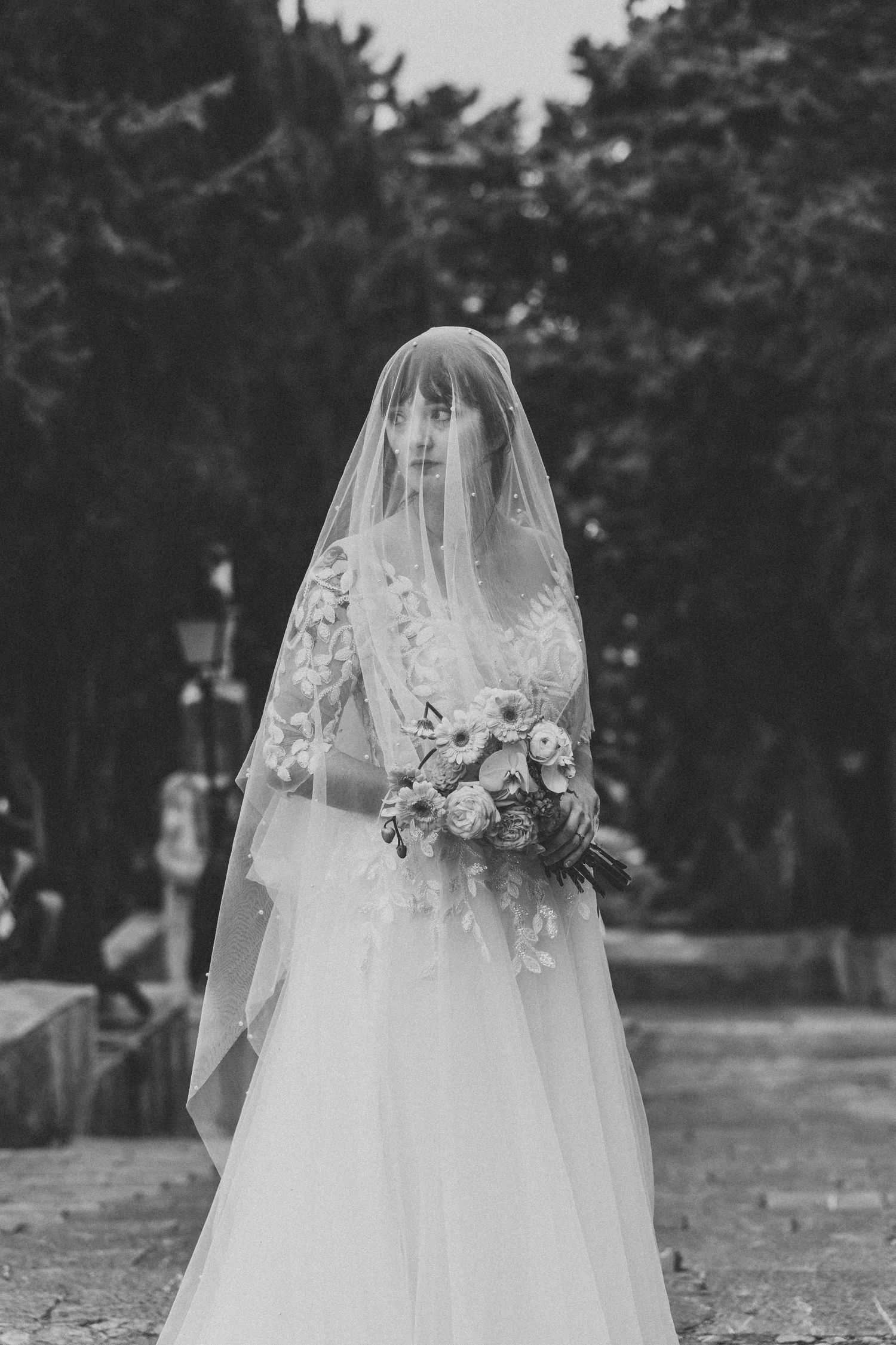 Black and white portrait of a bride in a flowing veil, holding a delicate bouquet with a serene expression.