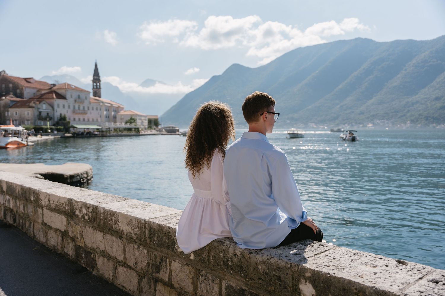 A couple sits on a stone wall overlooking a scenic Mediterranean coastal town, basking in the soft glow of the afternoon sun.