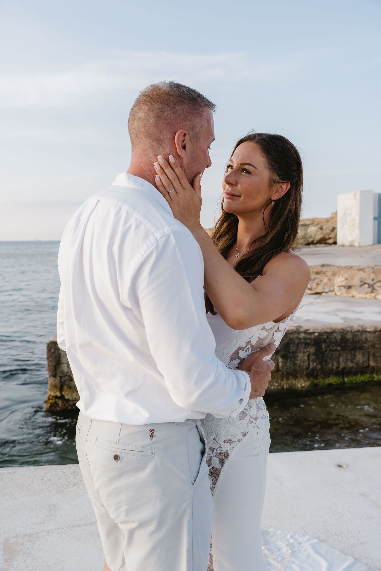 A newlywed couple in white embraces by the ocean at sunset, waves crashing gently behind them - romantic beach wedding in Malta.