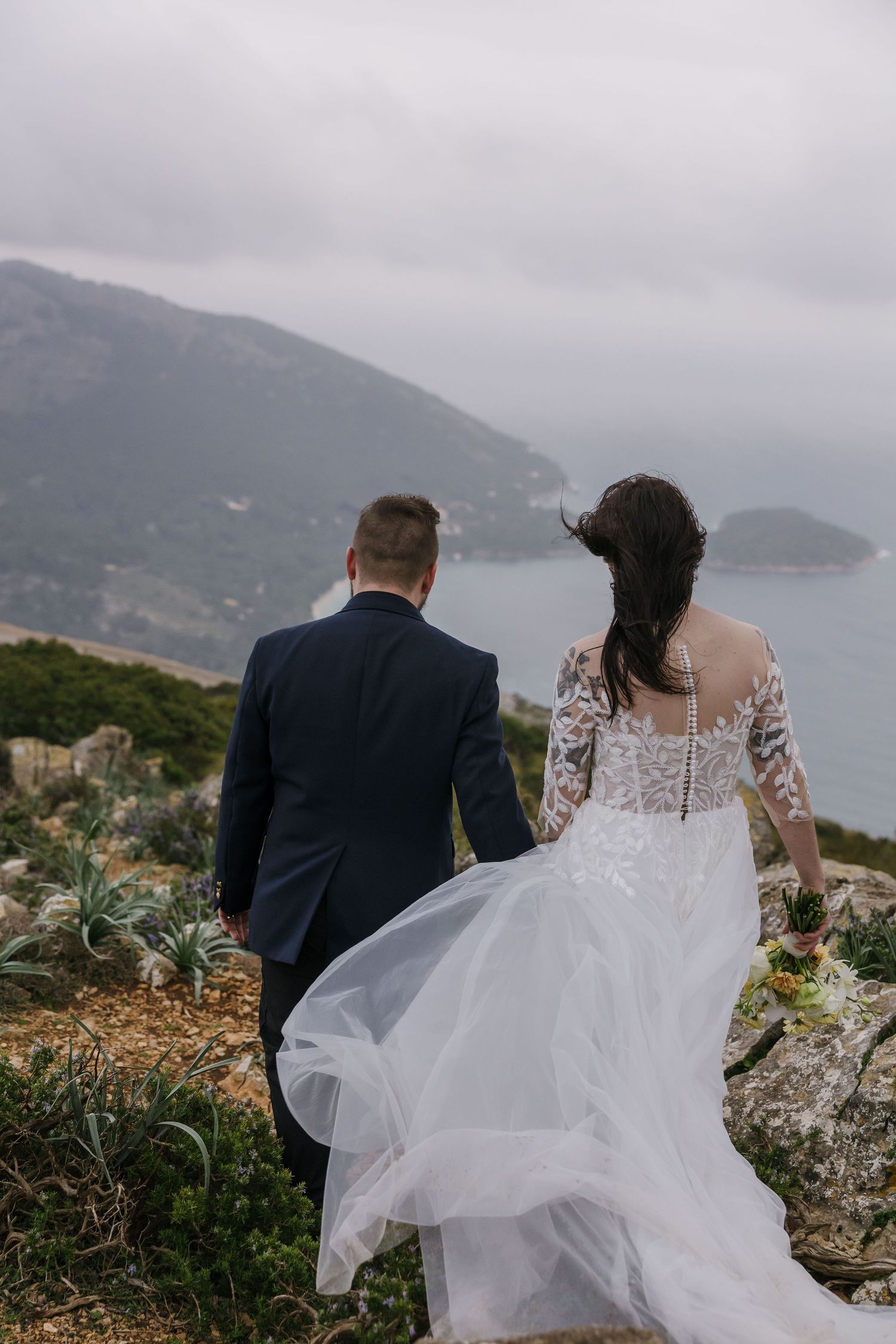 Bride and groom admire coastal view from mountaintop as bride’s dress flows in the wind - romantic elopement in Mallorca.
