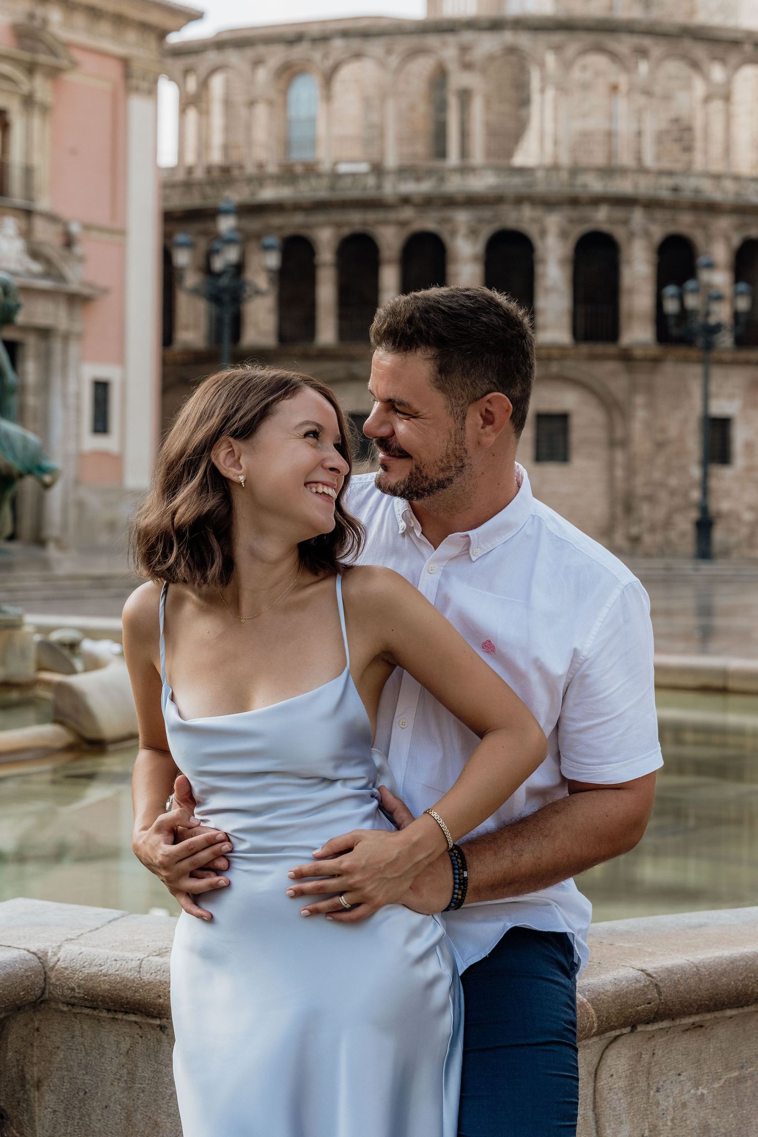 Romantic couple embraces by a historic fountain with European architecture in the background-timeless couples photography in Valencia, Spain