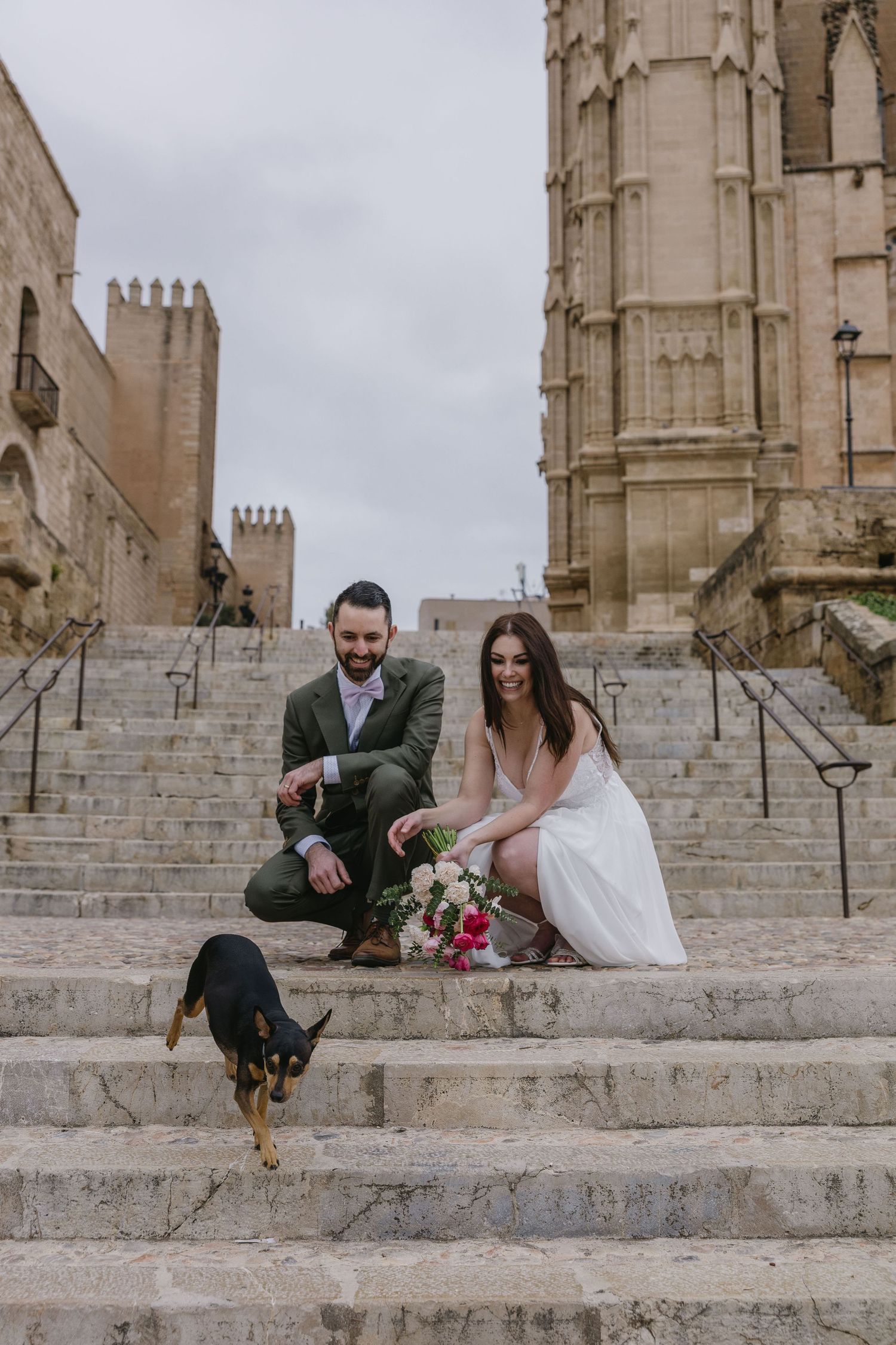 A newlywed couple poses with their dog on historic cathedral steps in Palma de Mallorca, blending romance with a heartfelt touch.