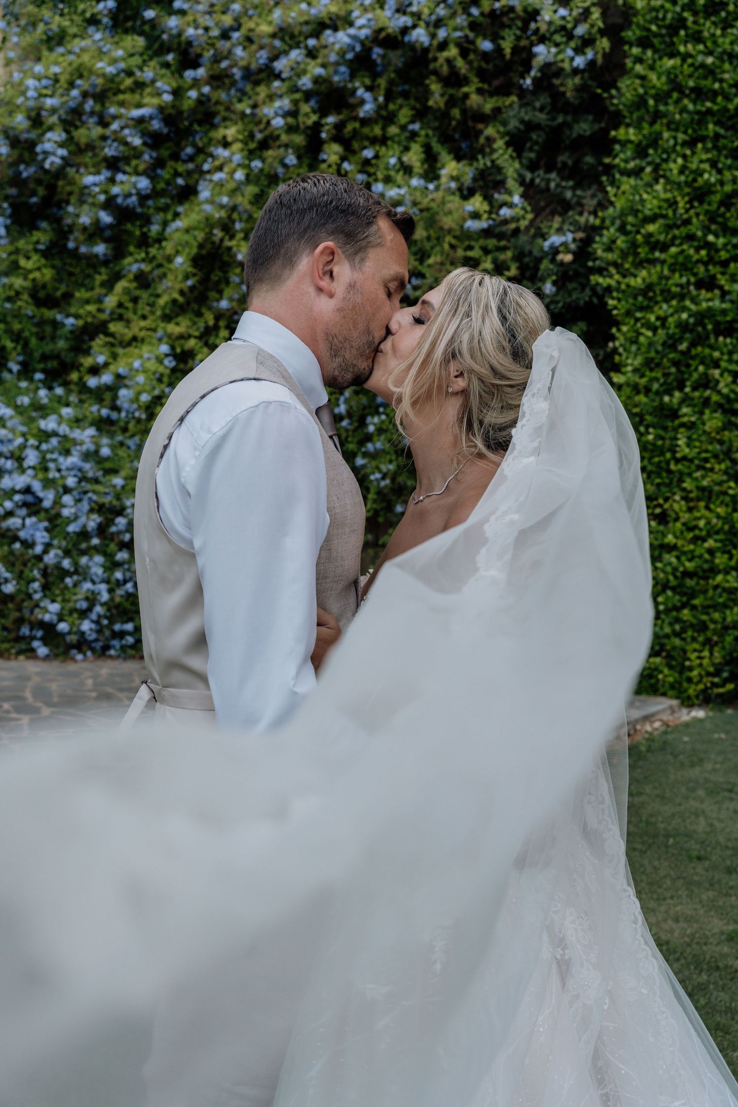 Bride and groom share a tender kiss as her veil flows in the breeze, framed by ivy-covered walls and soft evening light