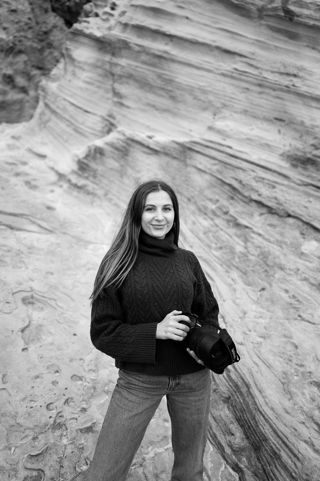 Photographer Kitija Banele standing against dramatic rock formations, camera in hand, capturing love stories with an artistic vision.