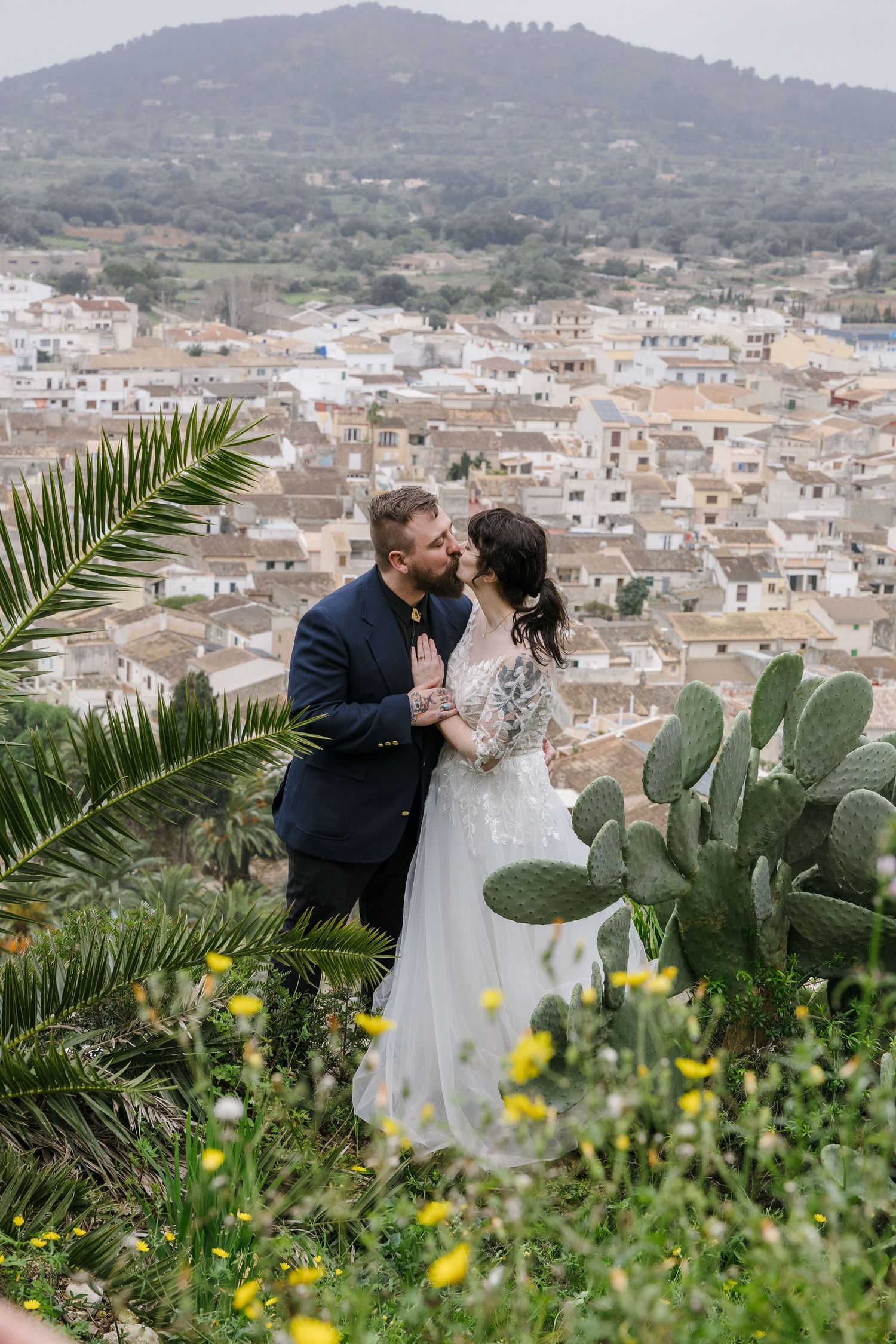 A newlywed couple embraces on a hillside in Mallorca, overlooking a sun-drenched Mediterranean village framed by cacti and palm fronds.