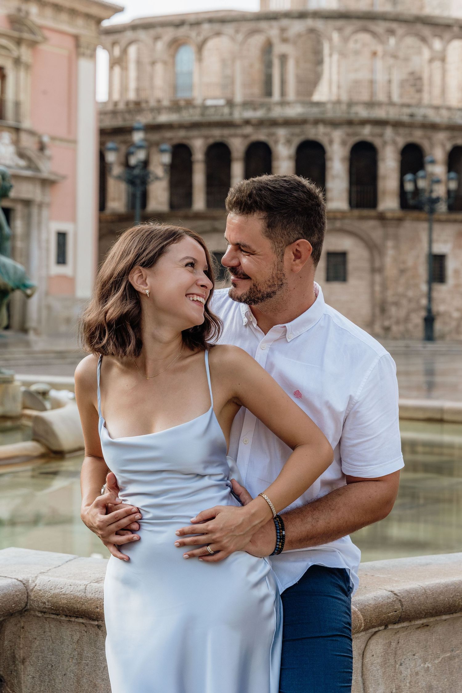 A couple laughs together by a historic fountain in Valencia, framed by elegant European architecture in the golden afternoon light.