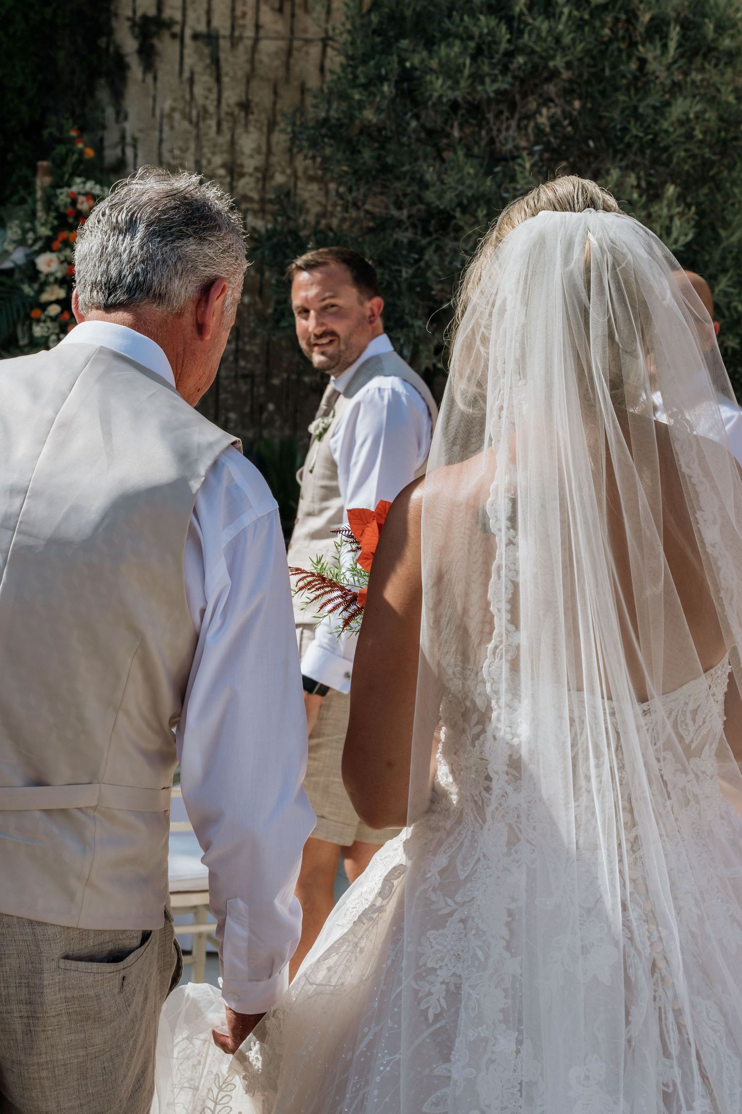 Bride walks hand in hand with loved ones during an intimate outdoor wedding, sunlight filtering through the trees.