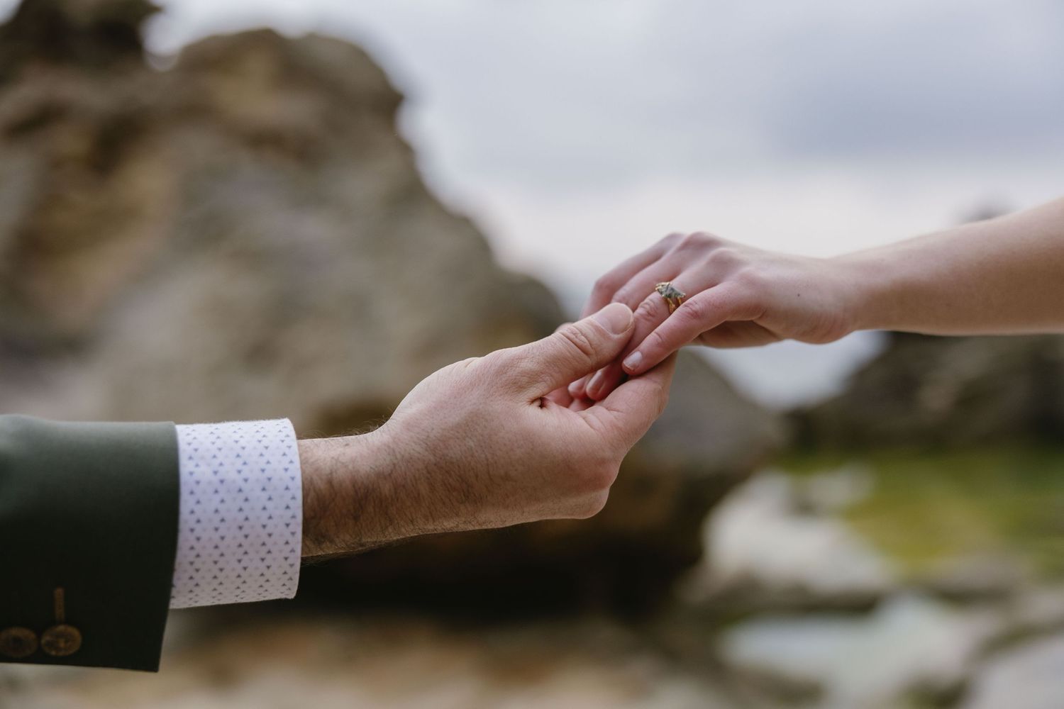 Close-up of hands gently exchanging wedding rings, set against a blurred coastal backdrop.