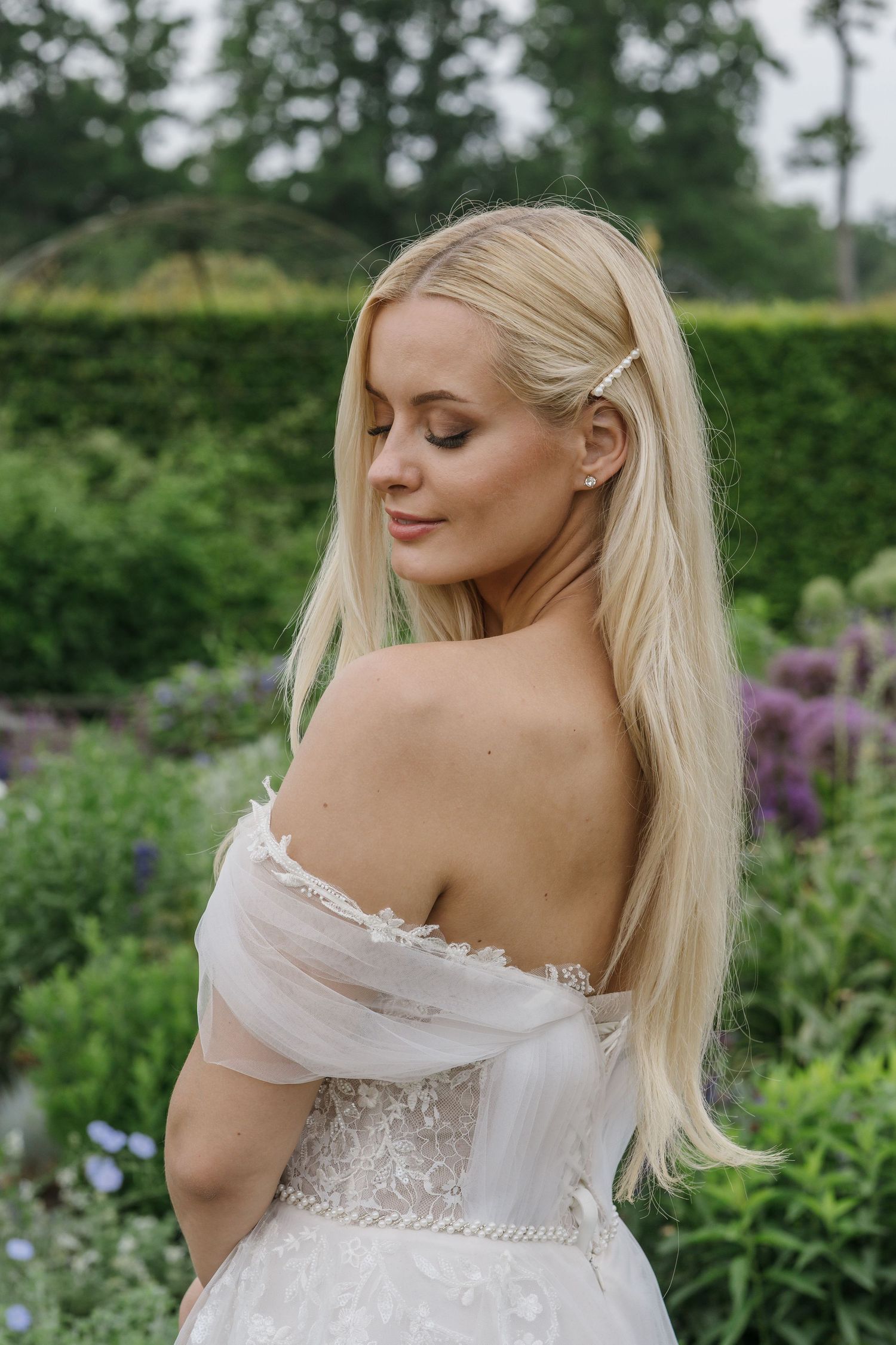 A bride in an off-shoulder wedding dress stands gracefully in a lush garden, surrounded by blooming purple flowers and soft greenery.