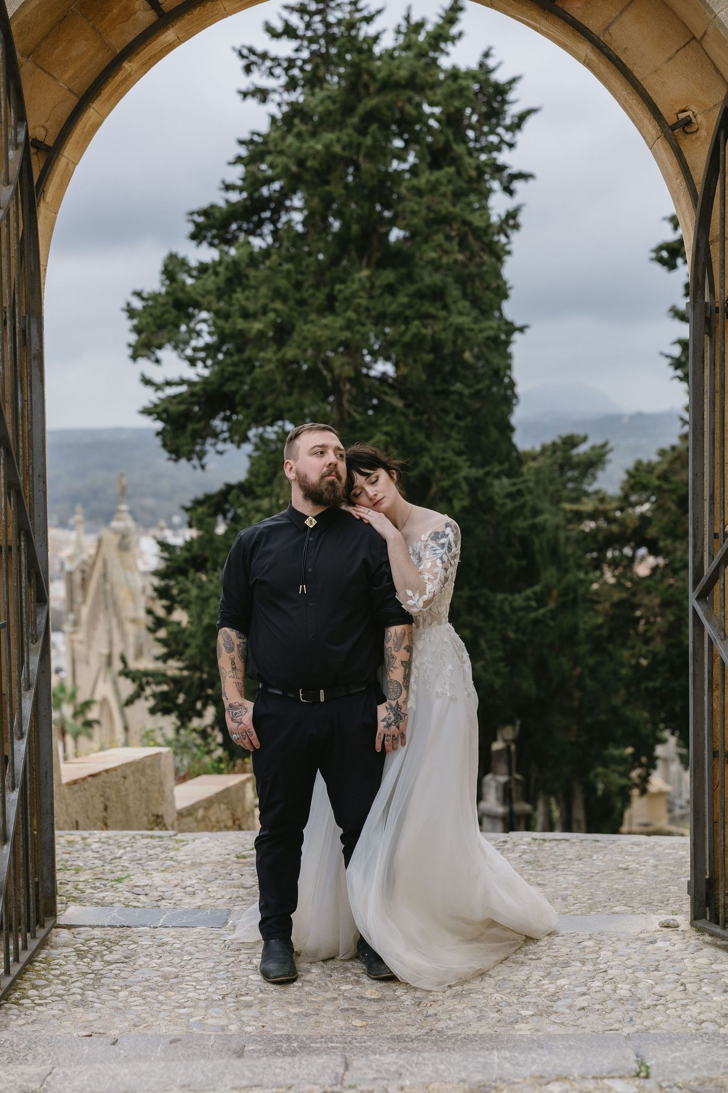 A newlywed couple stands beneath a rustic stone archway in Mallorca, with lush evergreen trees and a breathtaking European landscape behind