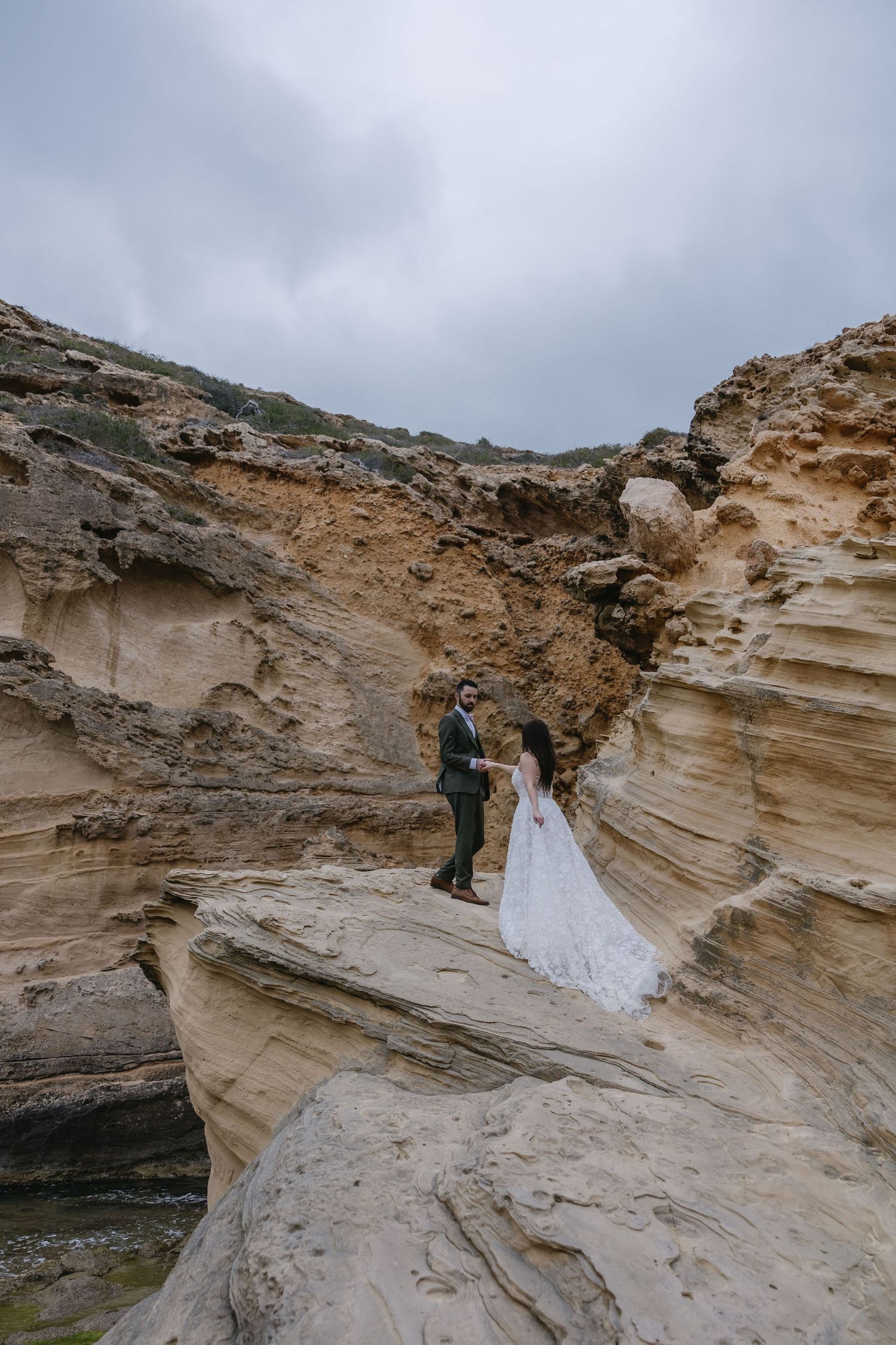 Bride and groom stand on dramatic rock formations under cloudy skies - adventurous elopement photography in Mallorca.