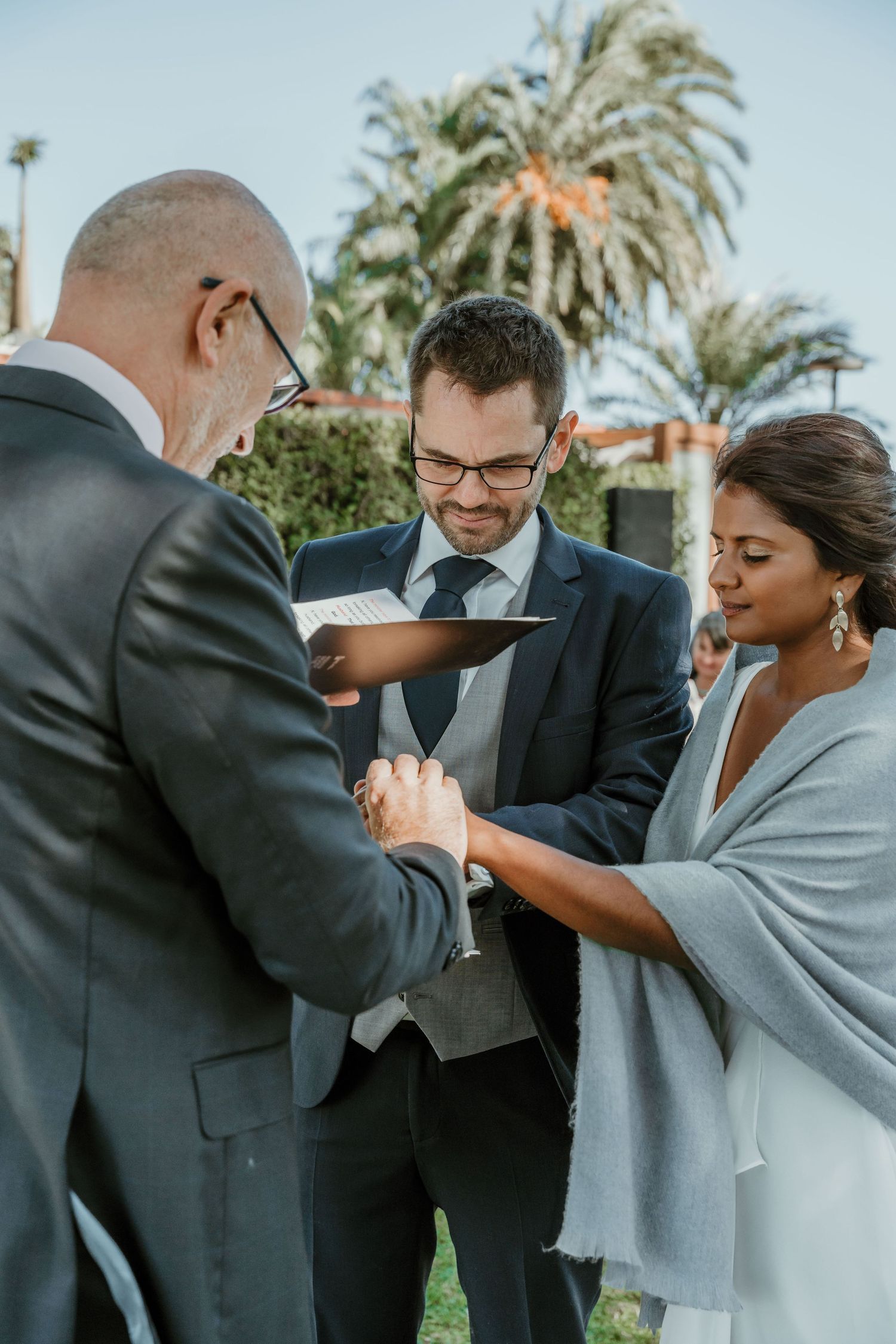 Bride and groom exchanging wedding rings in a scenic garden ceremony, officiated under palm trees with a warm and intimate ambiance.