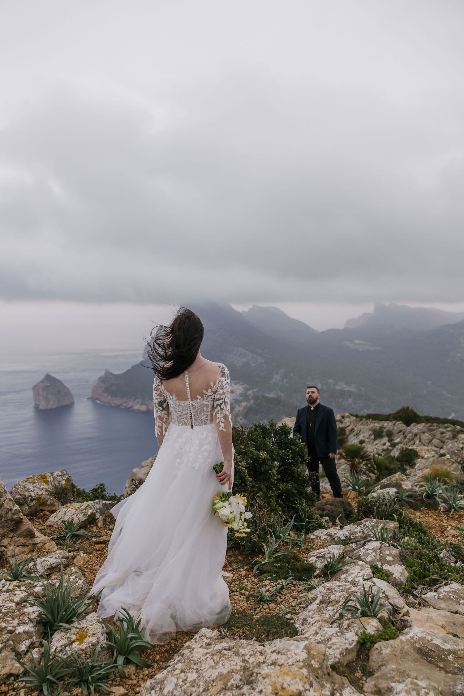 Elopement couple stands on rugged Mallorca cliffs, overlooking the dramatic coastline under moody skies - wild romance by the sea.