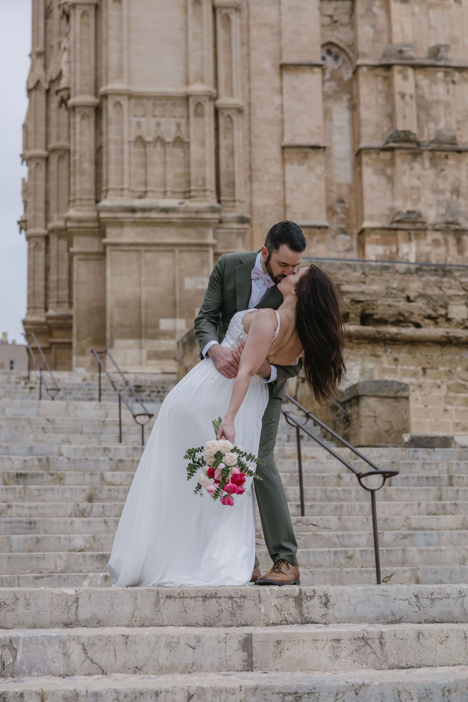 Newlyweds share a romantic embrace on the stone steps of a historic cathedral in Palma de Mallorca, framed by timeless European architecture