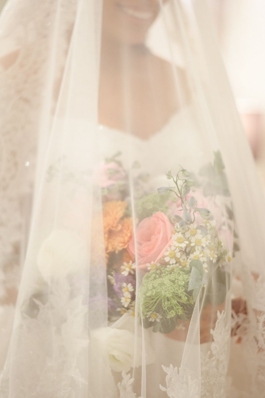 Bride holding colorful floral bouquet with roses and wildflowers, softly veiled in lace wedding veil.