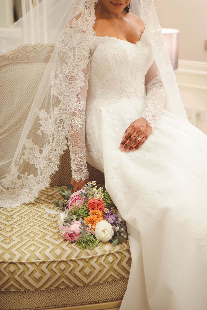 Bride in lace long-sleeve wedding gown and veil sits on ornate chair with colorful bridal bouquet beside her.