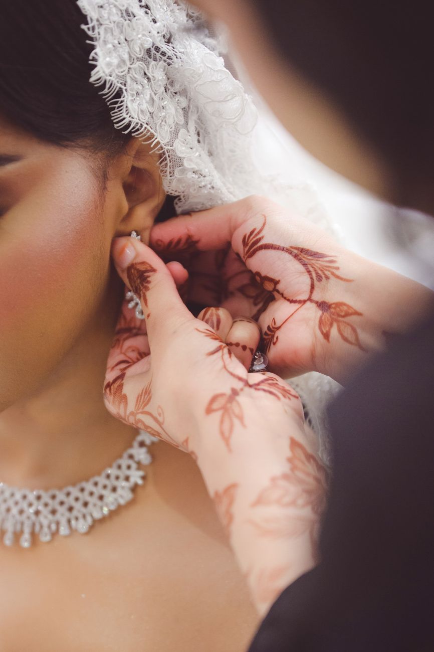 Bride with henna-adorned hands having earring placed, wearing pearl necklace and white lace veil on wedding day.