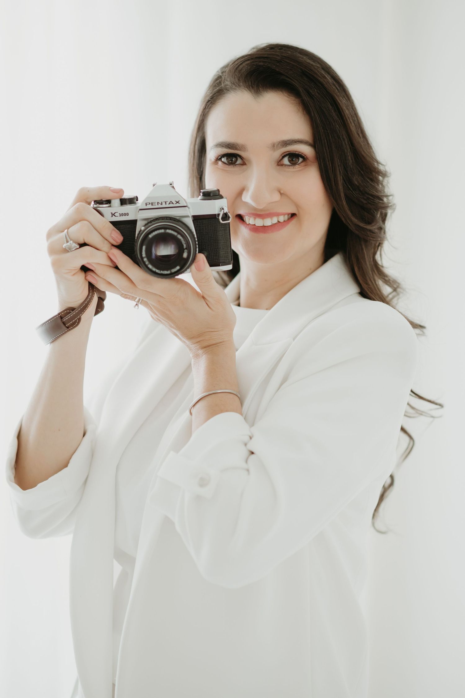 Smiling woman in white shirt holding a vintage Pentax film camera against a bright white background.