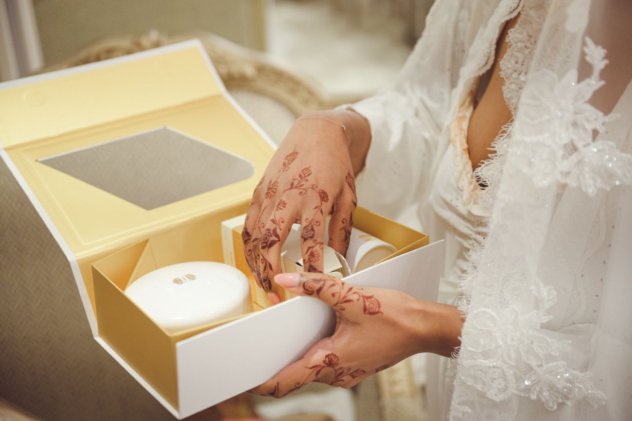 Bride with henna-decorated hands opening a yellow gift box in a white lace wedding dress.