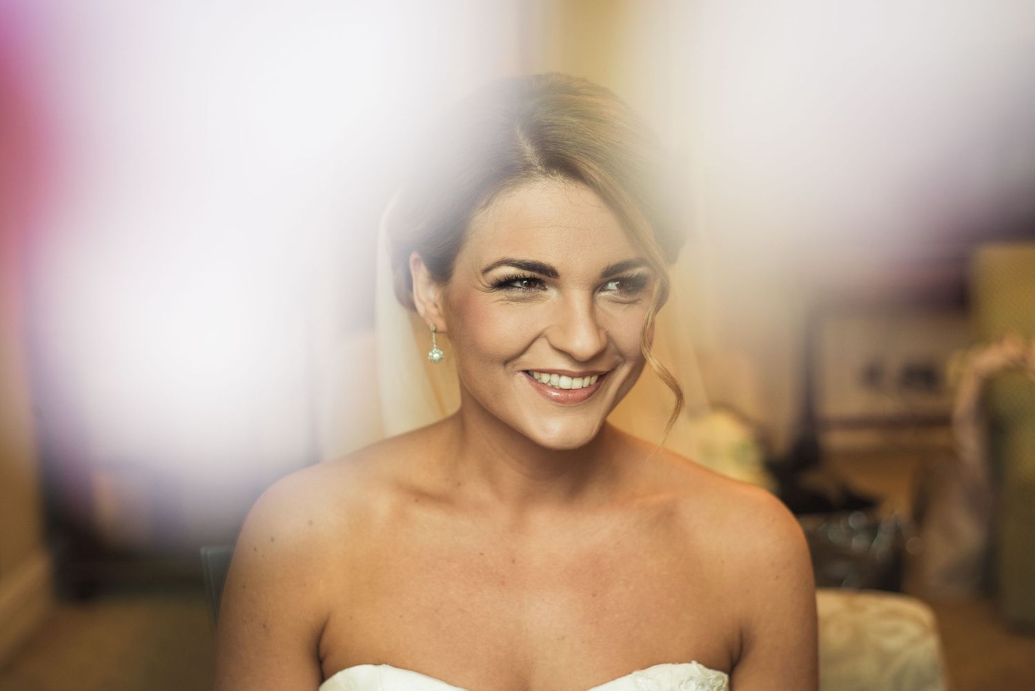 Smiling bride in strapless white dress with pearl earrings, glowing with joy on her wedding day.