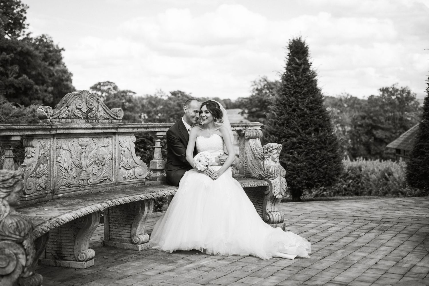 Black and white wedding photo of bride and groom posing on ornate stone bench in elegant garden setting.