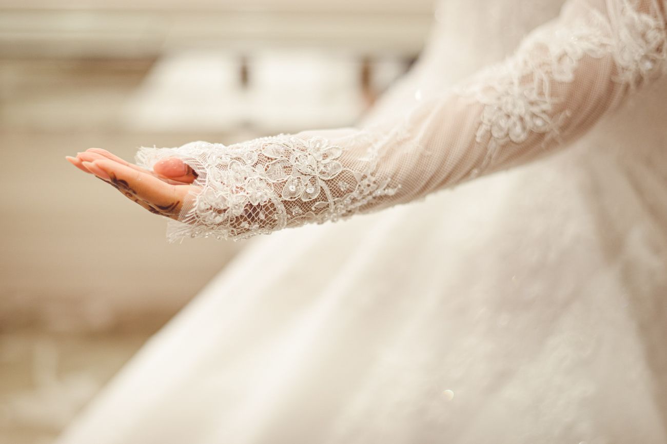 Close-up of a bride's lace gloved hand and elegant white wedding gown with intricate floral embroidery details.