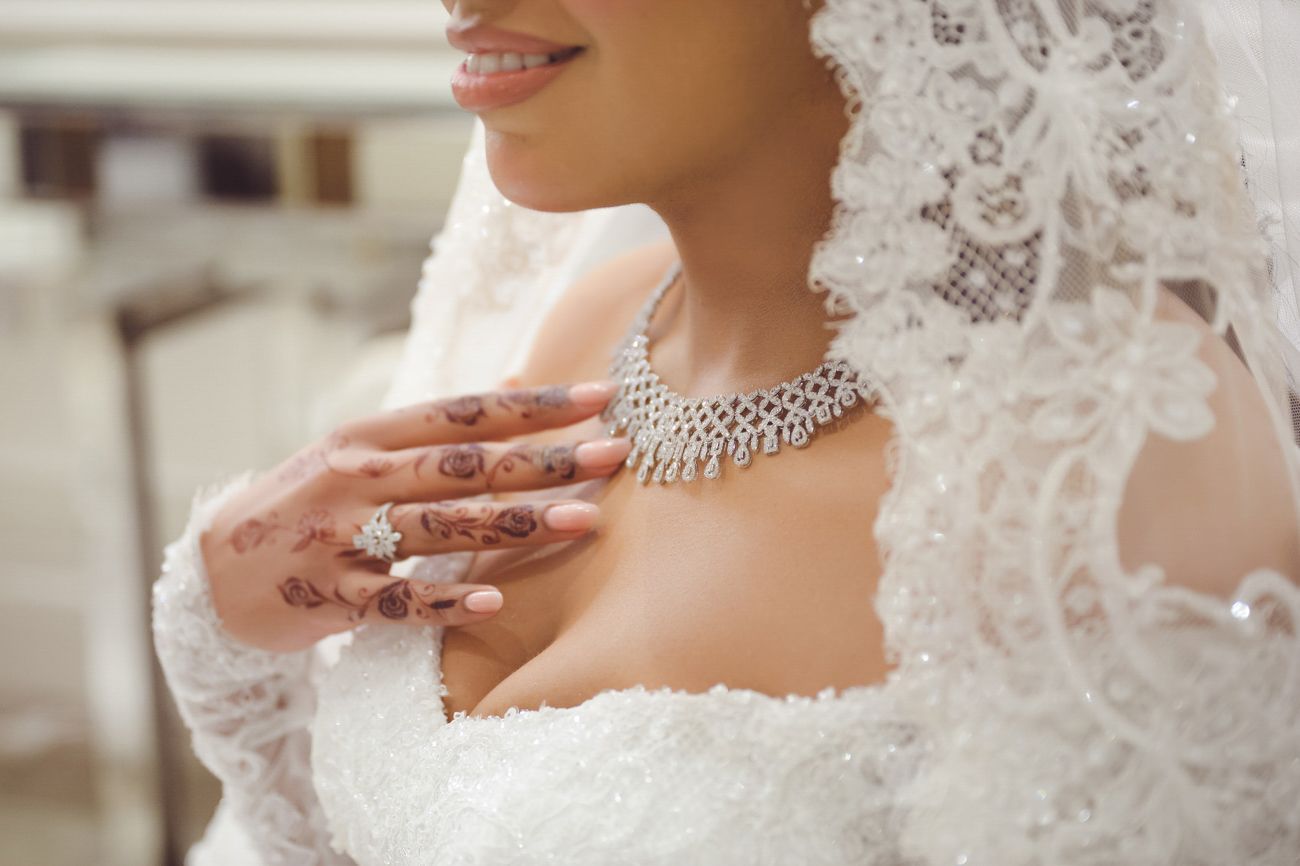 Bride in white lace wedding dress with henna hands, diamond necklace, and veil on her wedding day.