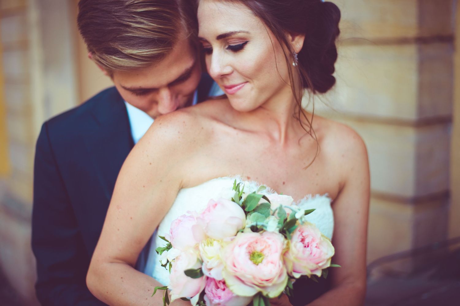 A bride holding pink peony bouquet leans into groom in navy suit during romantic outdoor wedding portrait.