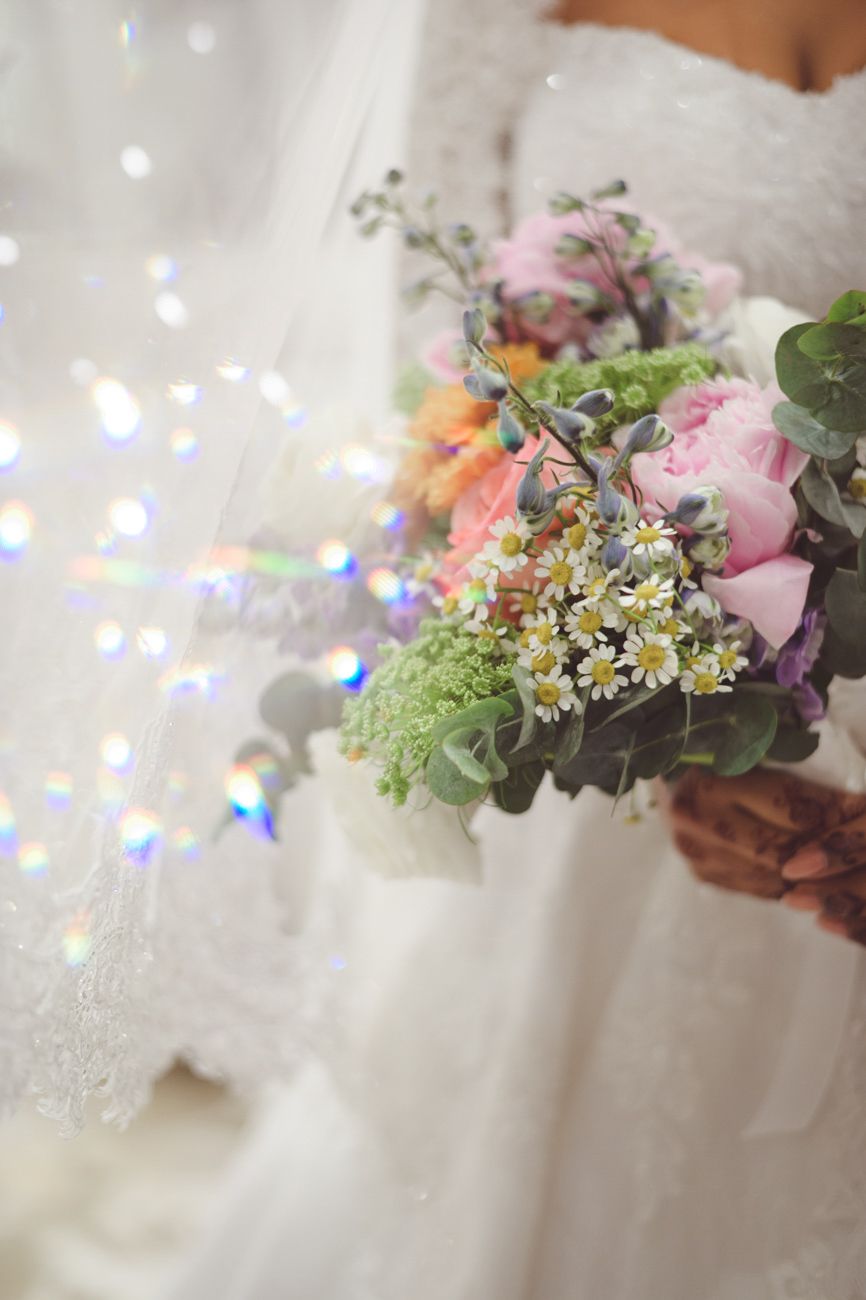 Bride holding a colorful wedding bouquet with roses, daisies, and greenery against a white lace dress.