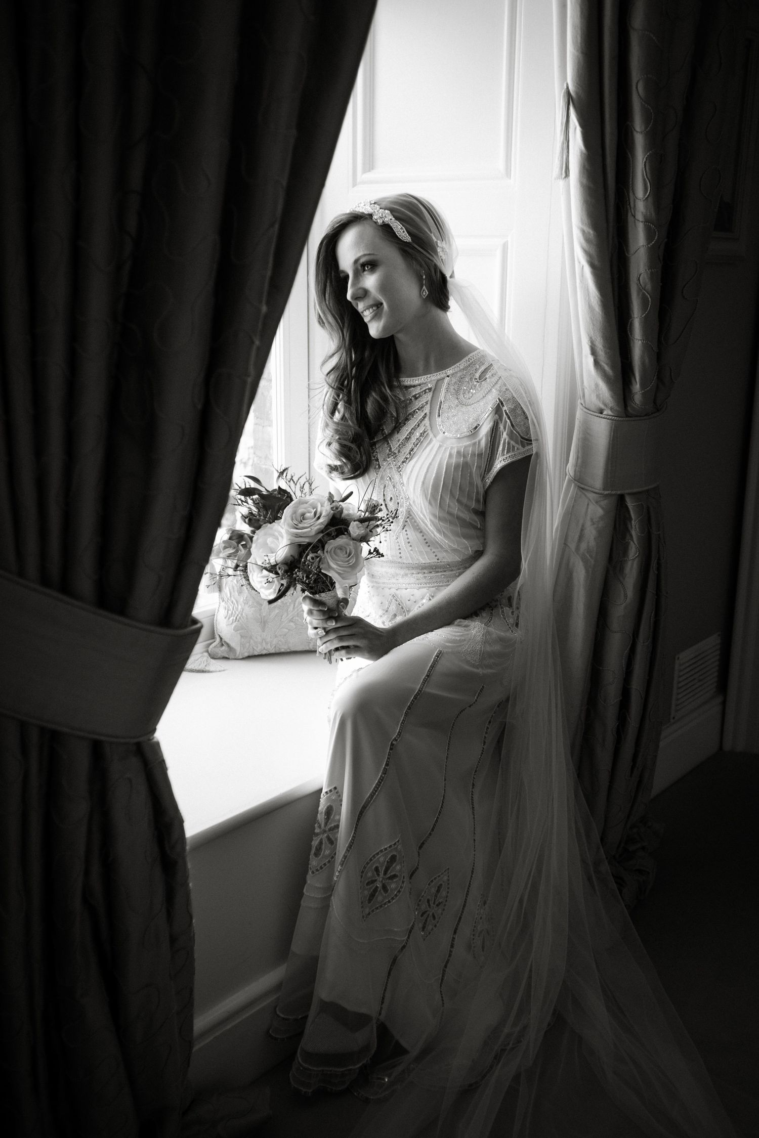 Black and white photo of a smiling bride in a lace gown holding a bouquet, seated by a sunlit window with draped curtains.