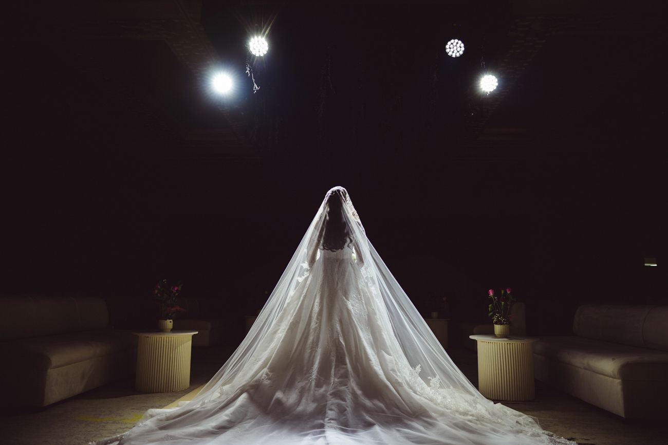 Bride in elegant white wedding gown with long veil, standing with back to camera under dramatic stage lighting.