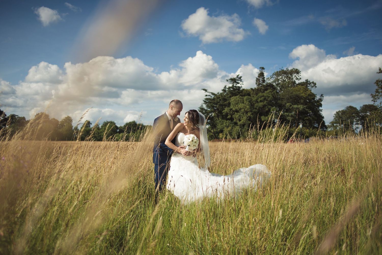 A bride and groom embrace in a golden meadow under a blue sky with fluffy clouds on their wedding day.