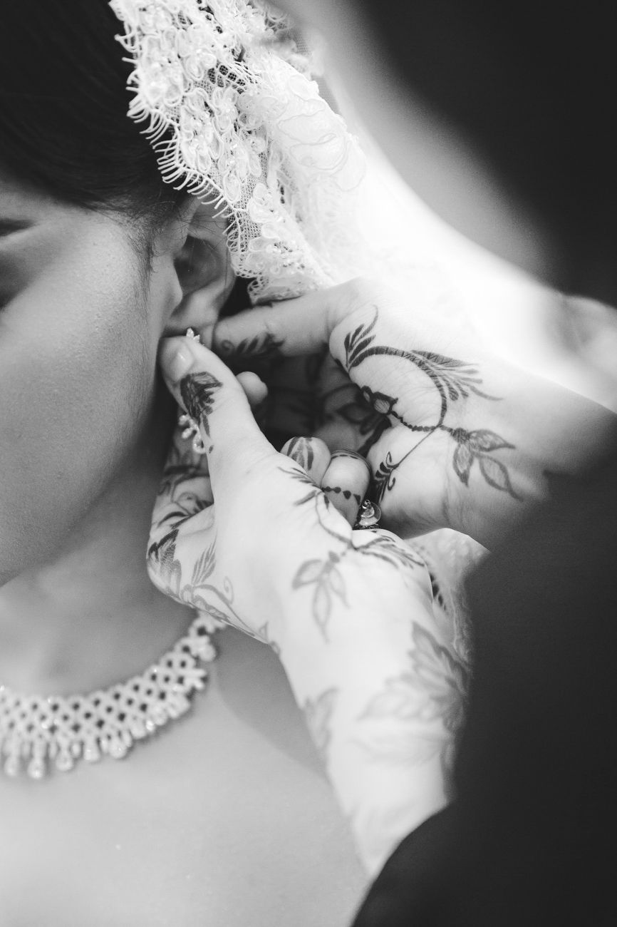 Black and white photo of a bride with henna-adorned hands adjusting her earring, wearing a pearl necklace and lace veil.