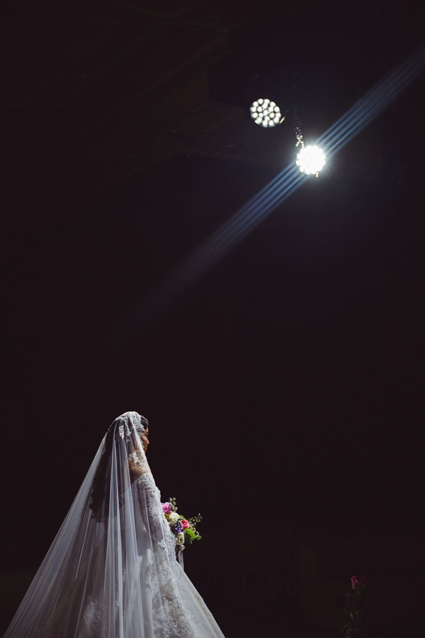 Bride in white lace gown and veil holds colorful bouquet under dramatic spotlight against dark background.