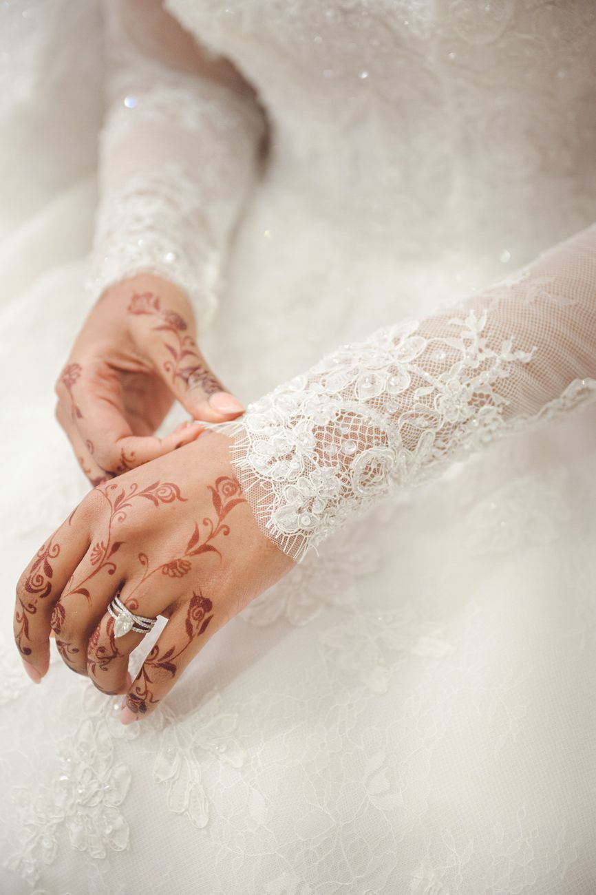 Bride's hands adorned with intricate henna designs and diamond ring, resting on white lace wedding dress.