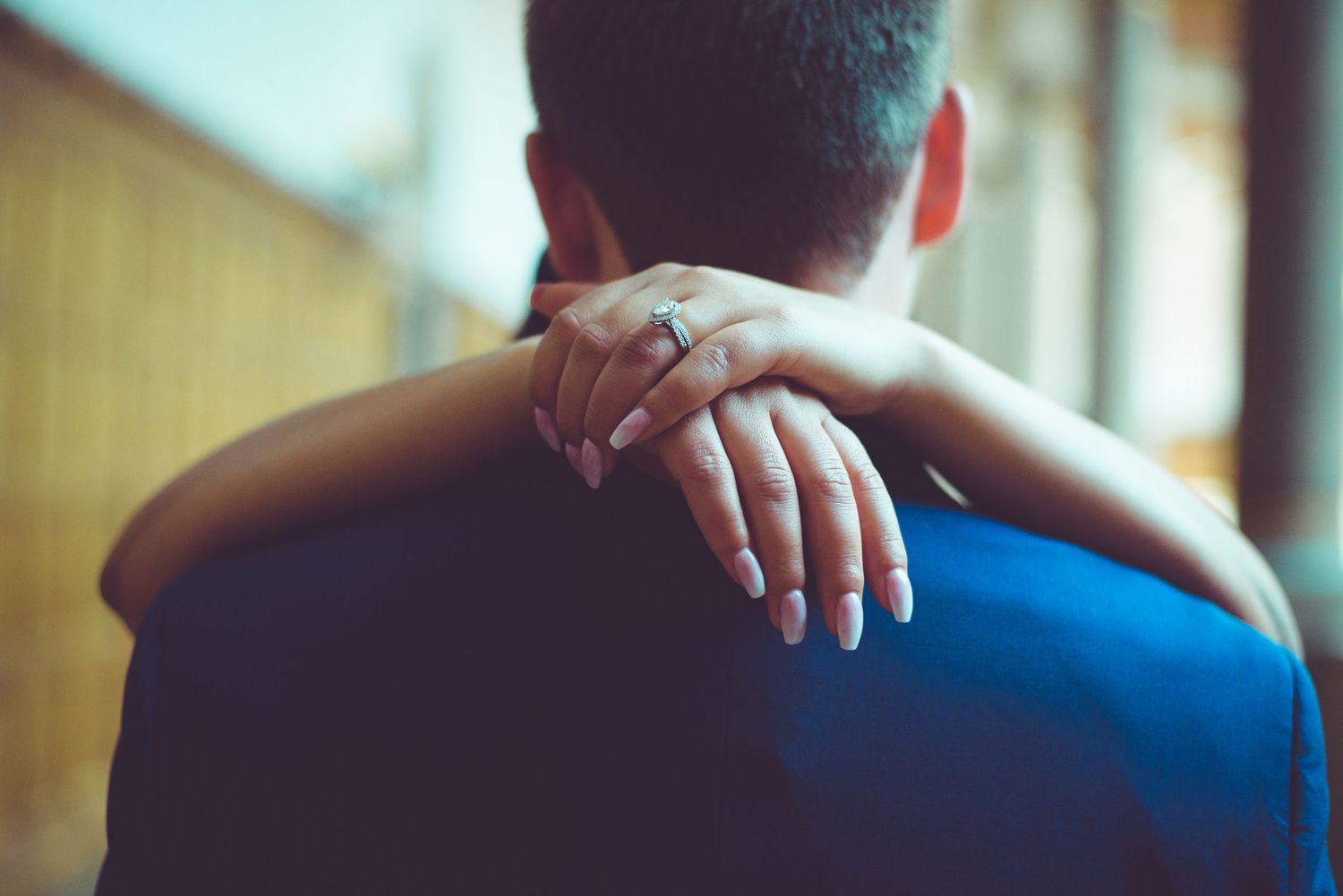 Woman with engagement ring embracing man from behind, couple sharing a romantic moment together.