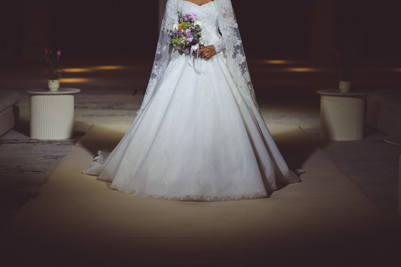 Bride in elegant long-sleeve white ball gown holding colorful bouquet, standing in softly lit venue.