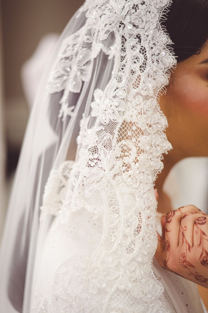 Bride wearing intricate white lace wedding veil with floral patterns, hands clasped in prayer.