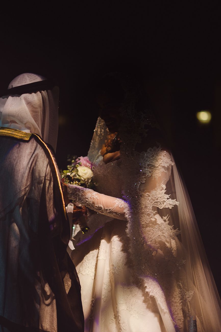 Bride in lace gown holding bouquet stands beside groom in traditional Arab attire during wedding ceremony.