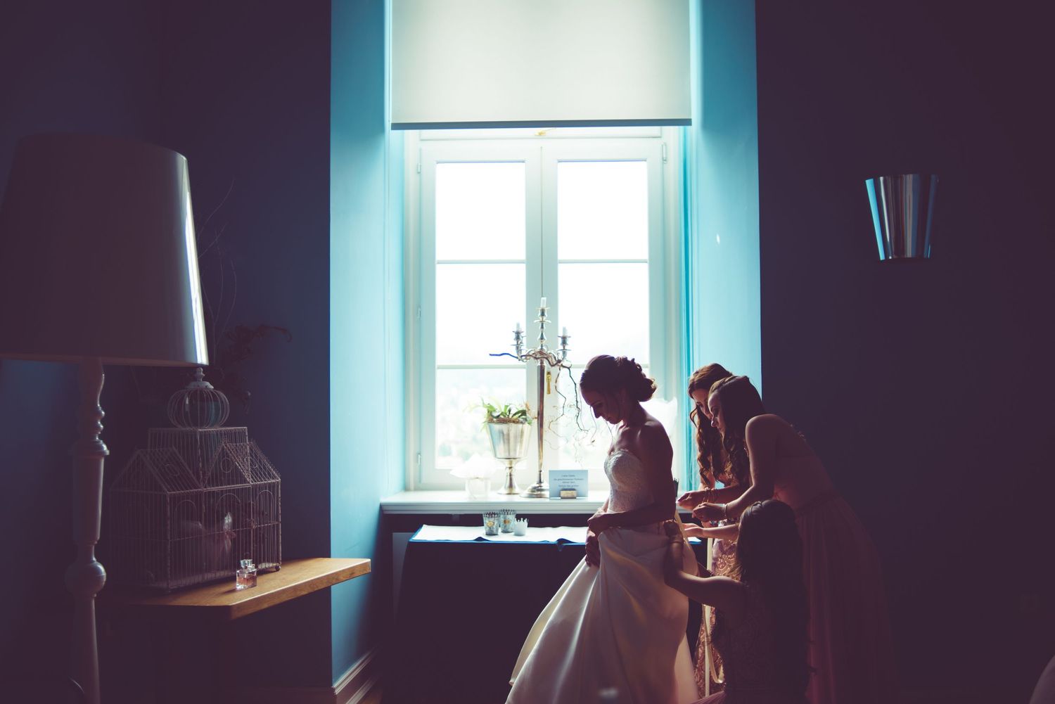 Bride in white gown silhouetted by window light in elegant blue room on wedding day.
