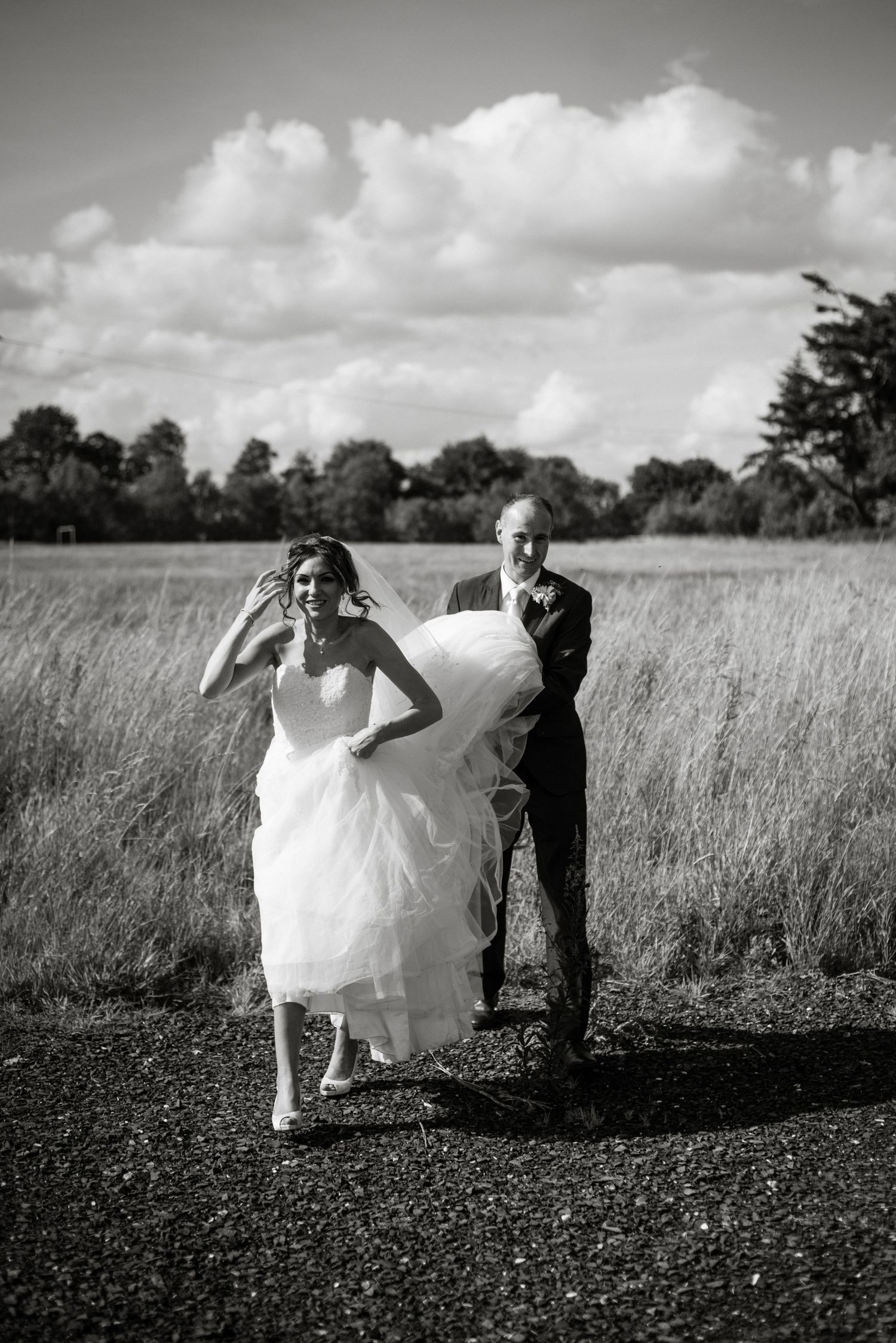 Black and white photo of a bride and groom running outdoors, man carrying wedding dress train, fields and clouds behind.